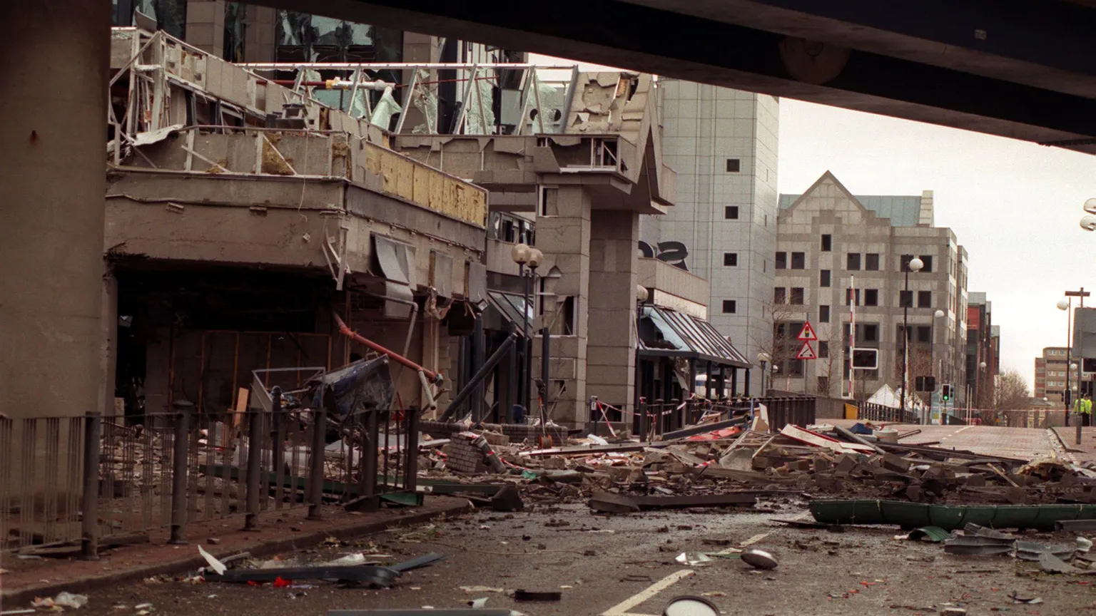  A photo from 1996 of debris strewn across the scene of the IRA bomb blast in London's Docklands. It is a big grey building. There is rubble across the road.