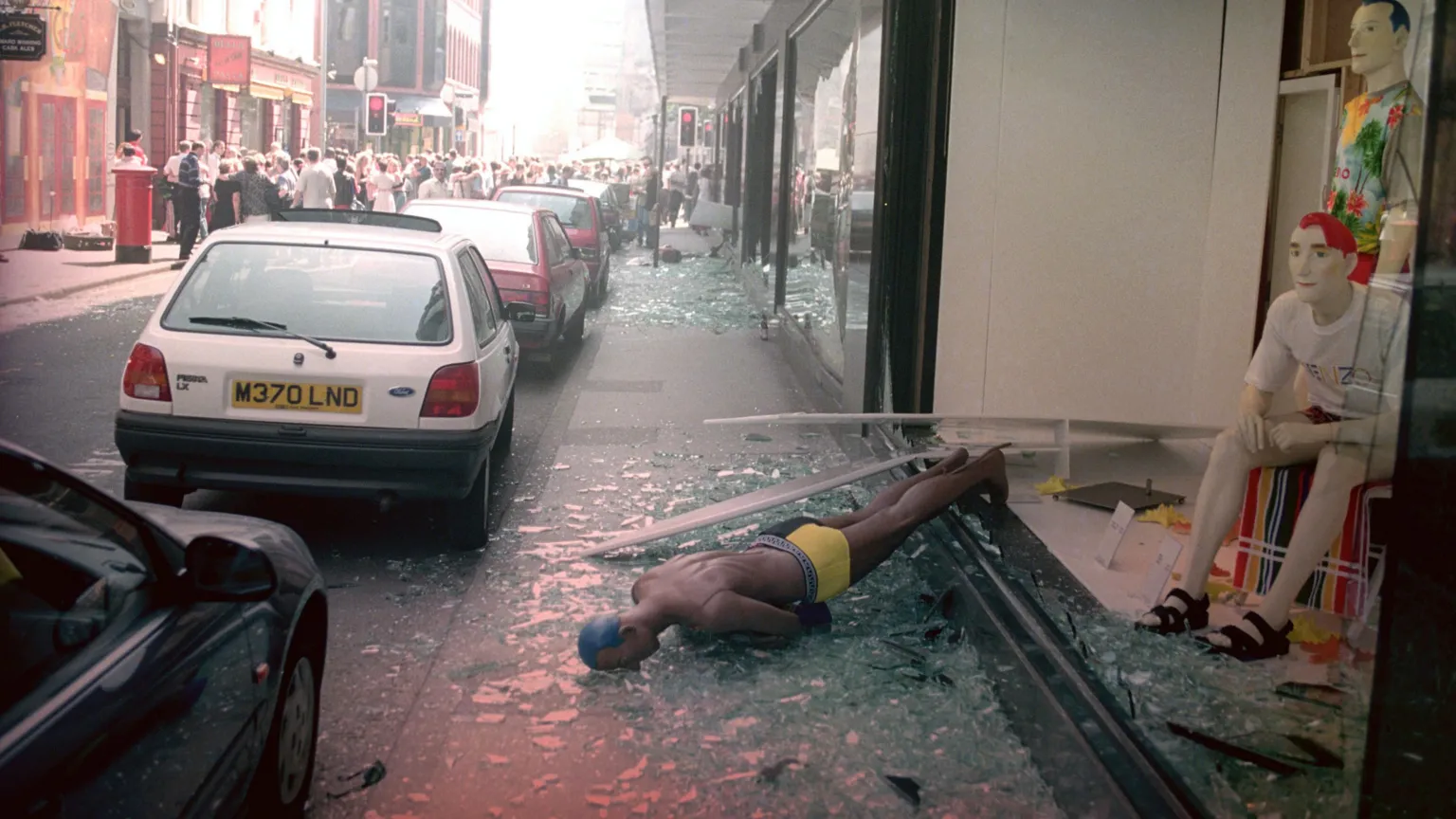  A mannequin blown through a shop window, lies on the pavement following a bomb attack at the Arndale Centre. Broken glass can be seen on the pavement. Cars are parked on the street and a crowd of people are standing further down the street.