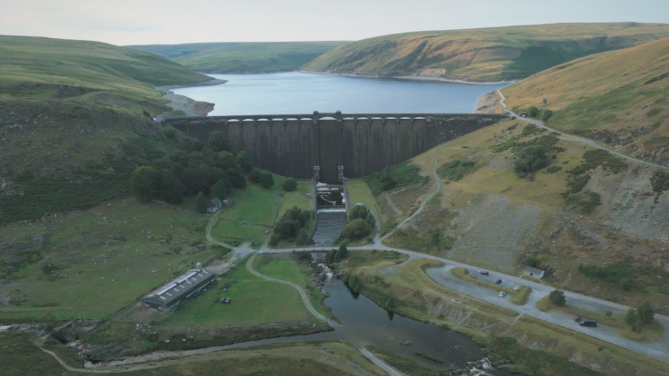 Llun wedi'i dynnu o uchder gan ddrôn o gronfa ddŵr Claerwen. Mae'n dangos yr argae gyda'r gronfa tu ôl iddi. Mae modd gweld afon a chaeau gwyrdd.