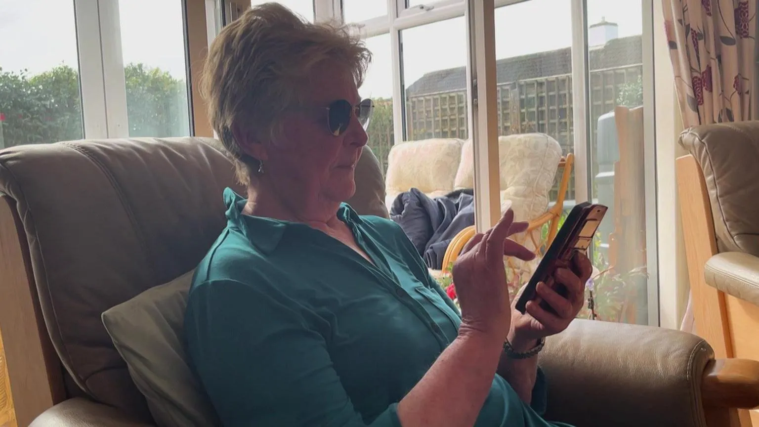 An elderly woman called Ruth Goodman sits on a chair in her living room while looking at her mobile phone. She has glasses on and is wearing a teal blouse.