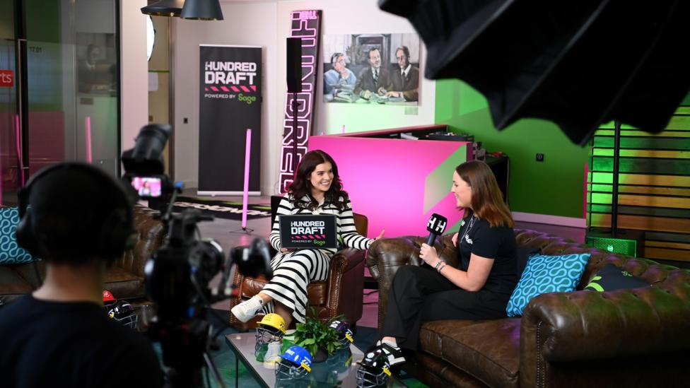 An interview being conducted in a room at The Shard, there is a camera man in the foreground and two women sat on sofas in the middle. 'The Hundred' draft branding is in the background 