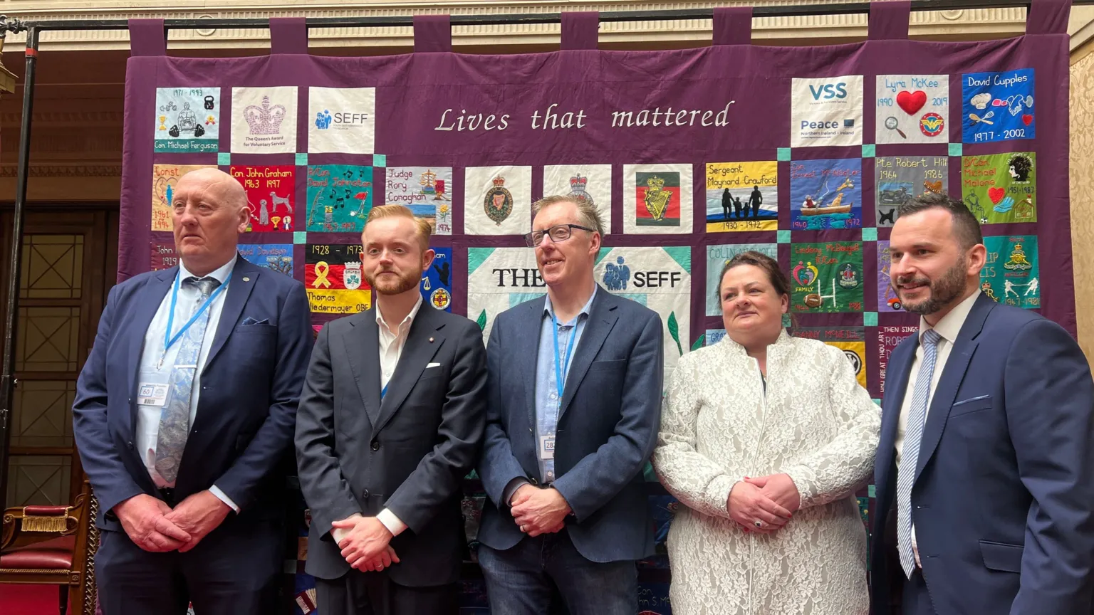 Five people standing in a row. Four men are wearing blue suits and a woman is wearing a cream jacket.