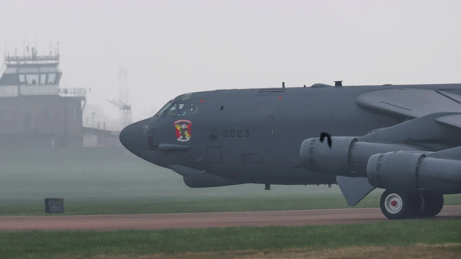 A large black B-52 bomber. It is a long sleek looking plane with a sticker on the side of the cockpit saying 'Bomber Barons'. It is coming down the runway on a misty day.