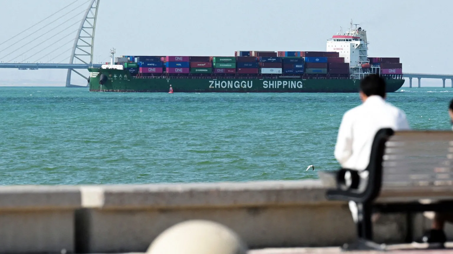 AFP via A person sits on a bench next to the sea. In the distance a container ship heads towards a port in Kuwait City.