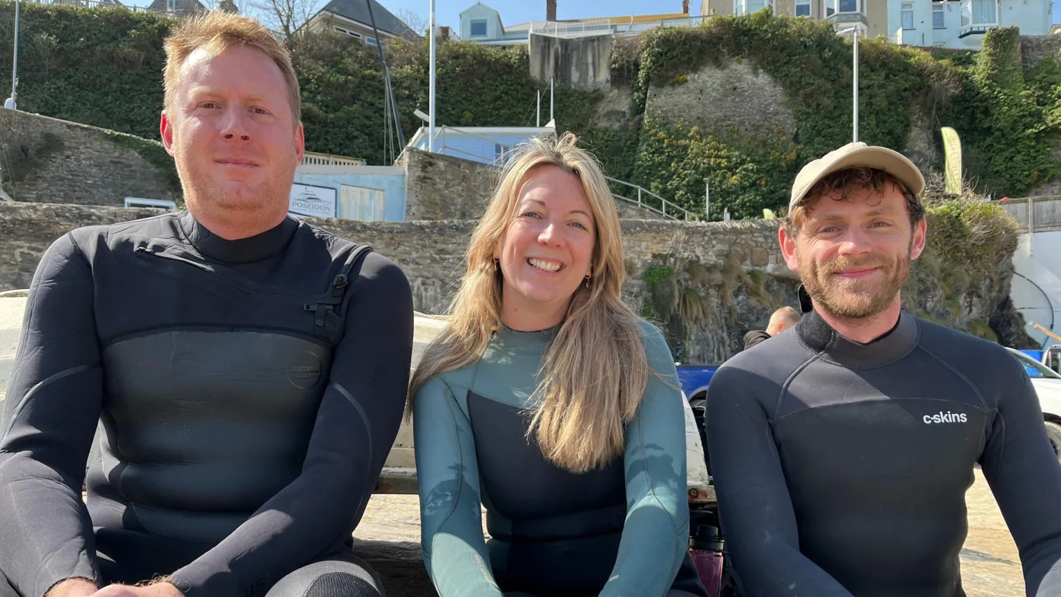 Three people are sat on a stone wall outside. The person in the middle is a woman the other two are men. They appear to be in their early 40's. They're wearing wetsuits. The woman and the man on the left, look happy and relaxed the man on the right appears more anxious. All three have their hands clasped in front of them either resting on their knees or between their legs. It's a sunny day and there are stone houses further up the hill behind them.