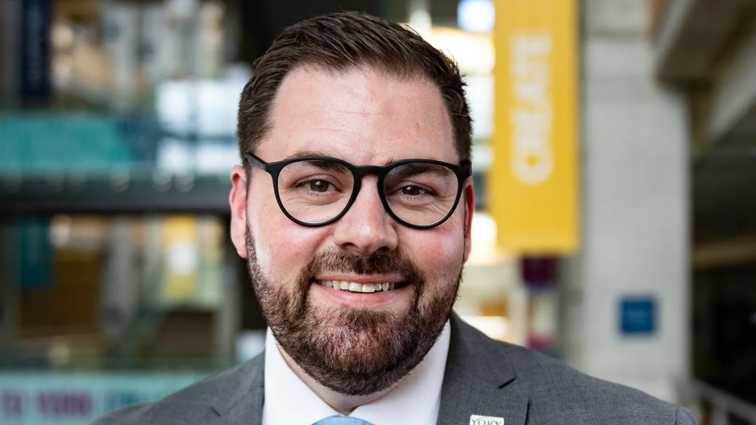Dan Rose/We Are Awesome Ken Merry is smiling at the camera, wearing a grey suit and blue tie. He has a beard and black, round-framed glasses.