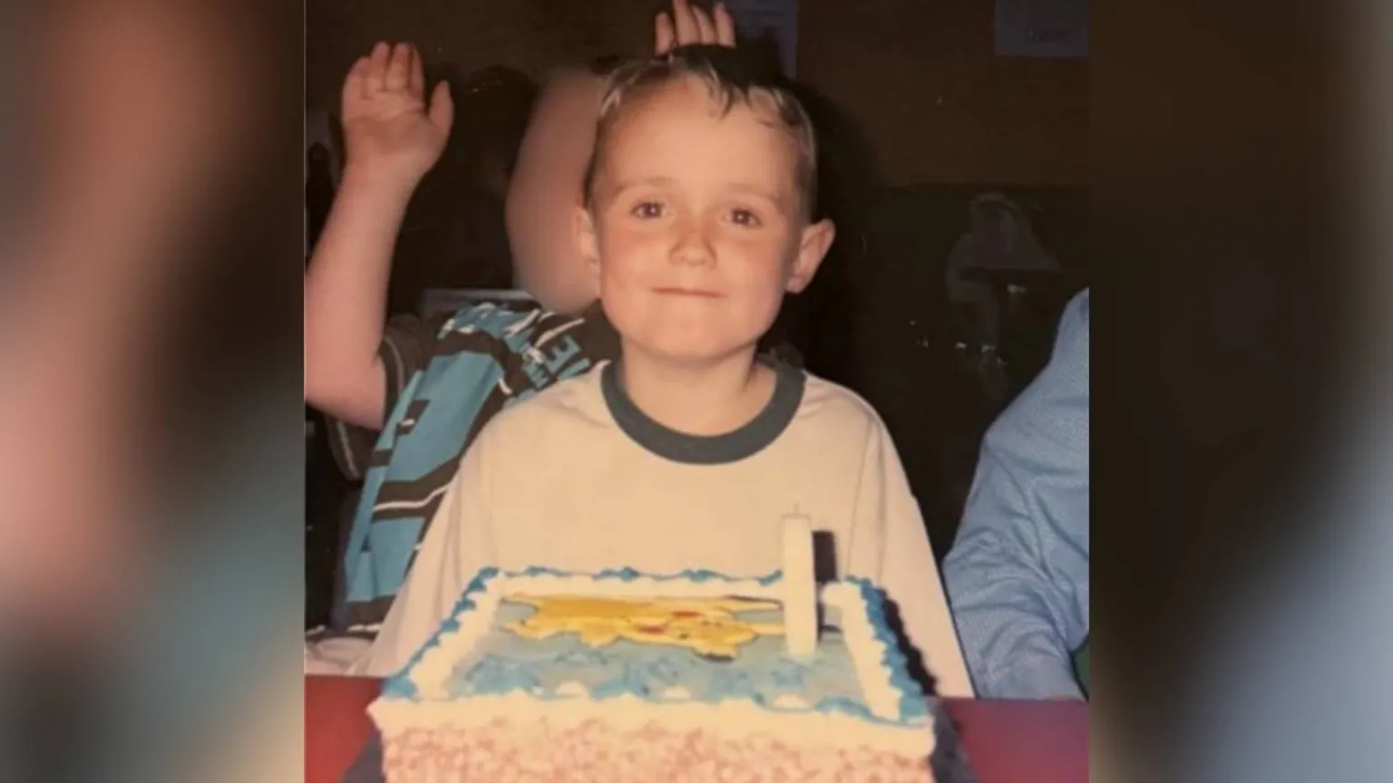 Ben Cole Edwards Photograph of Ben Cole Edwards sat in front of his pokemon birthday cake at the forefront. He has brown eyes and brown hair and smiles at the camera. A young boy can be seen behind him, however his face is blurred. Ben wears a white t-shirt with a navy trim collar. 