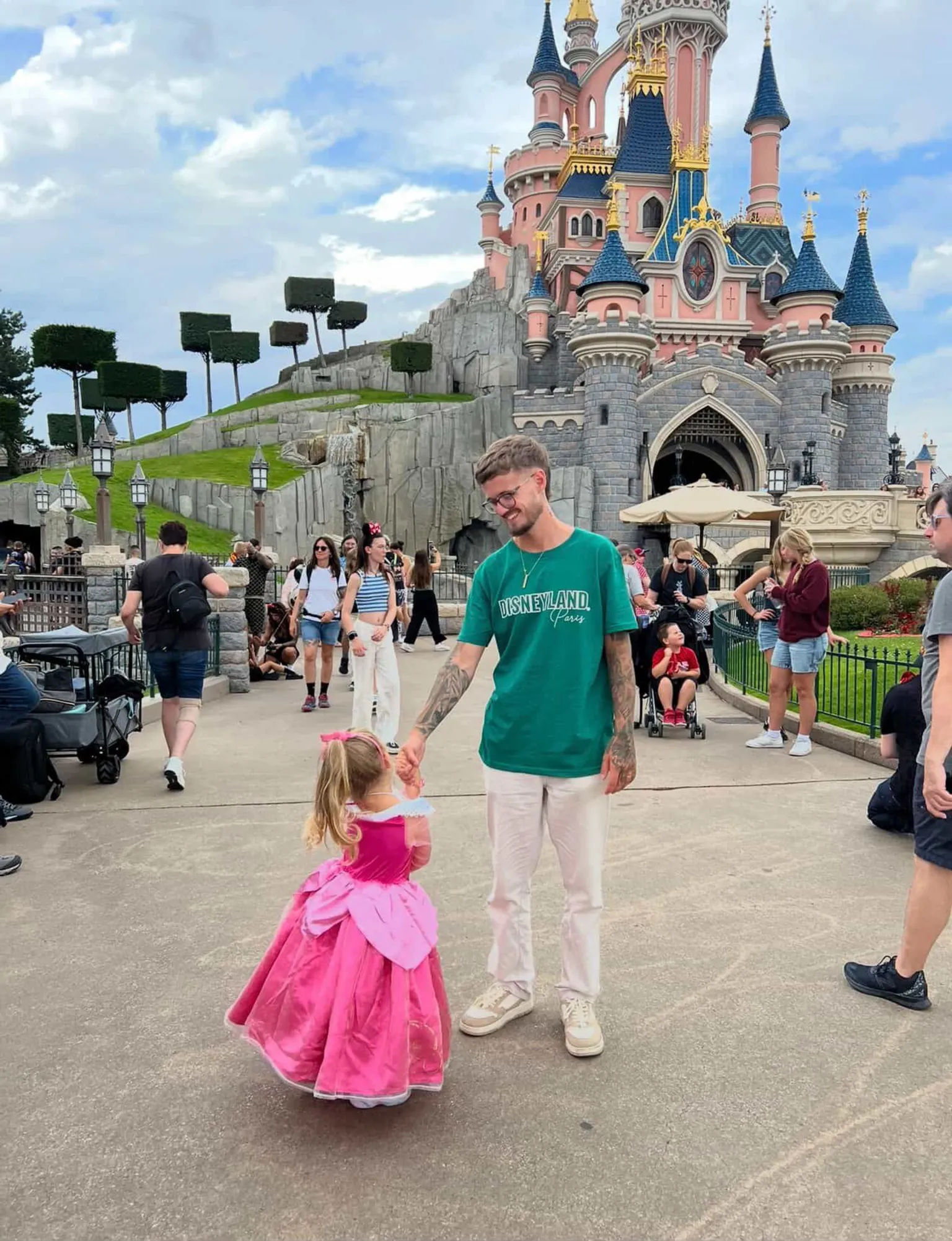 Ben Cole Edwards Photograph of Ben Cole Edwards, who is 30 years old and has brown hair, holding his daughters hand. He and his daughter stand in front of a castle at Disneyland Paris. His daughter wears a pink princess dress and has blonde hair in a ponytail. 