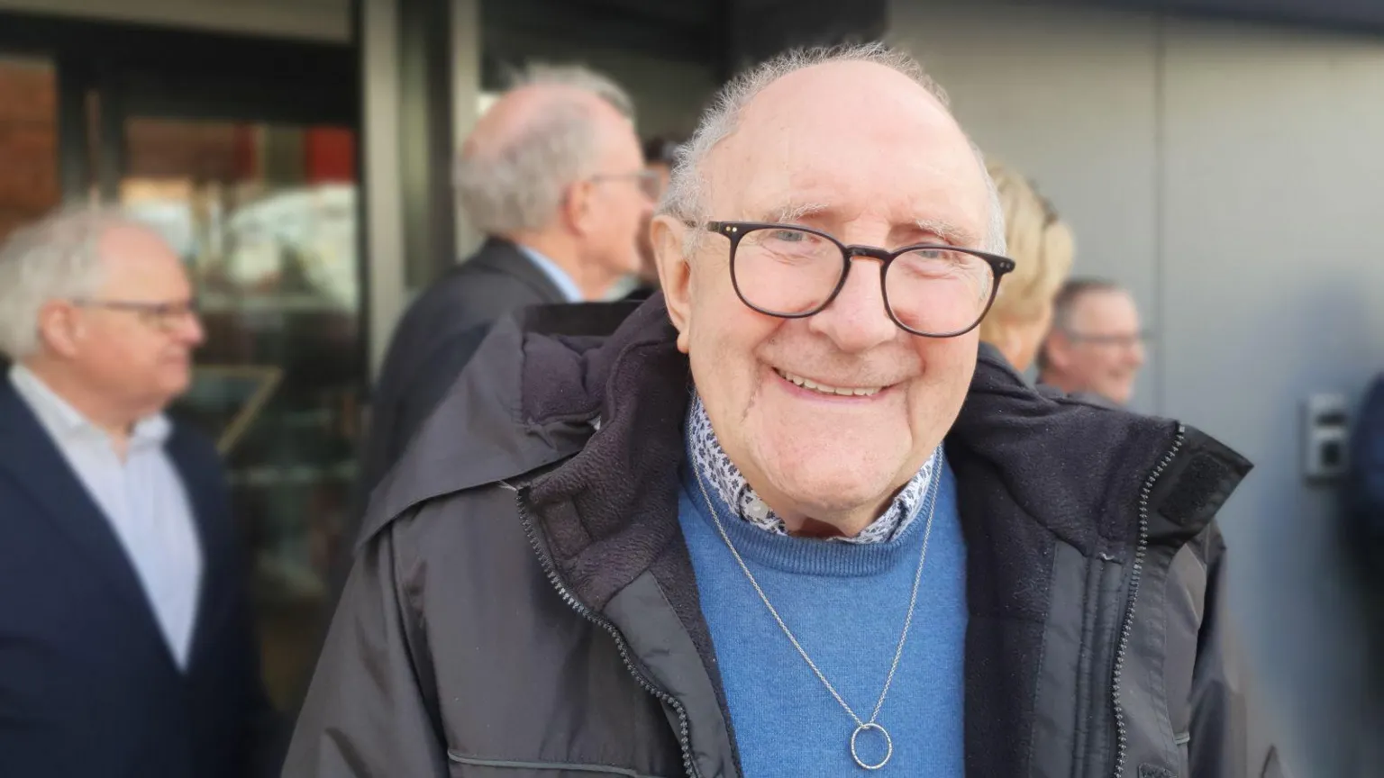 A man with dark rimmed glasses, wispy white hair, a blue jumper, silver necklace and black over coat smiles at the camera. 