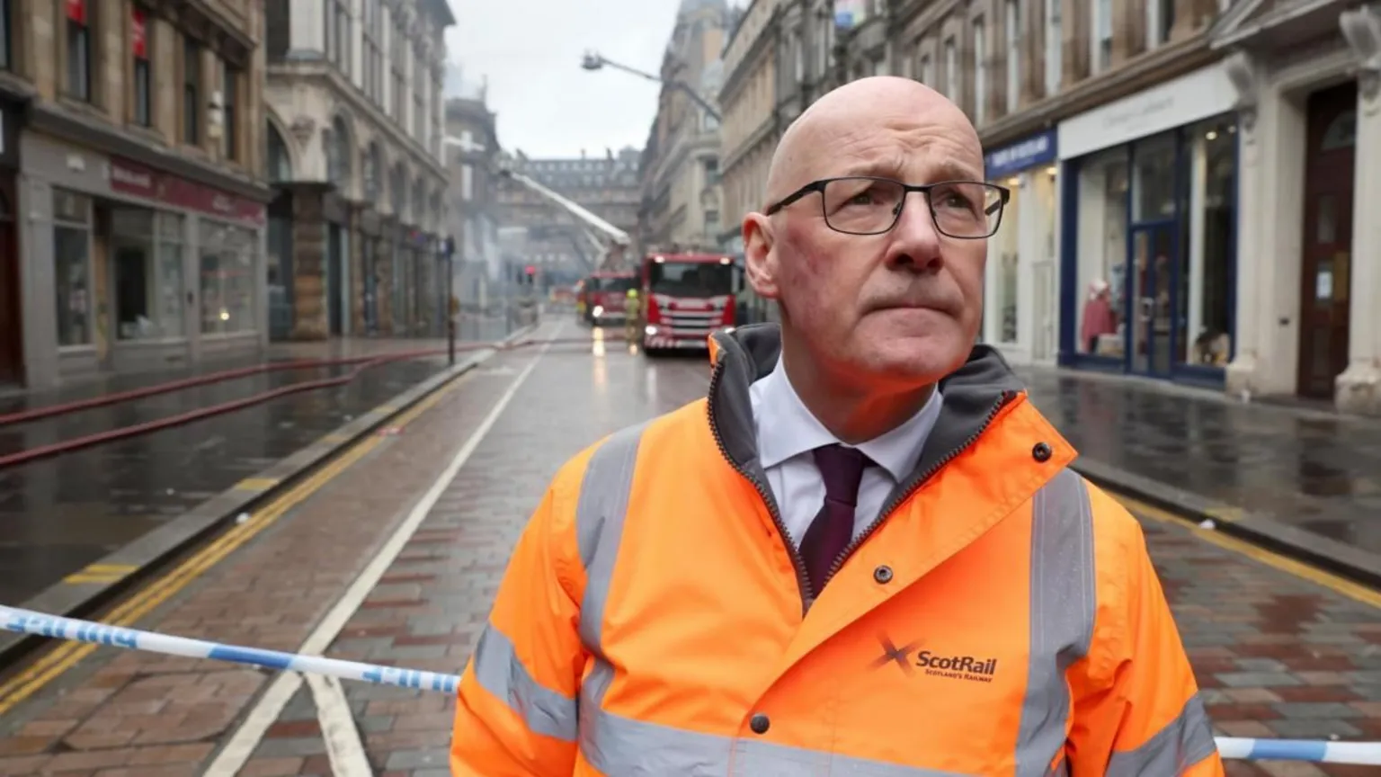  Swinney wearing a bright orange ScotRail high‑visibility jacket stands in front of an emergency cordon on a wet city street, with fire crews and fire engines working in the background amid smoke