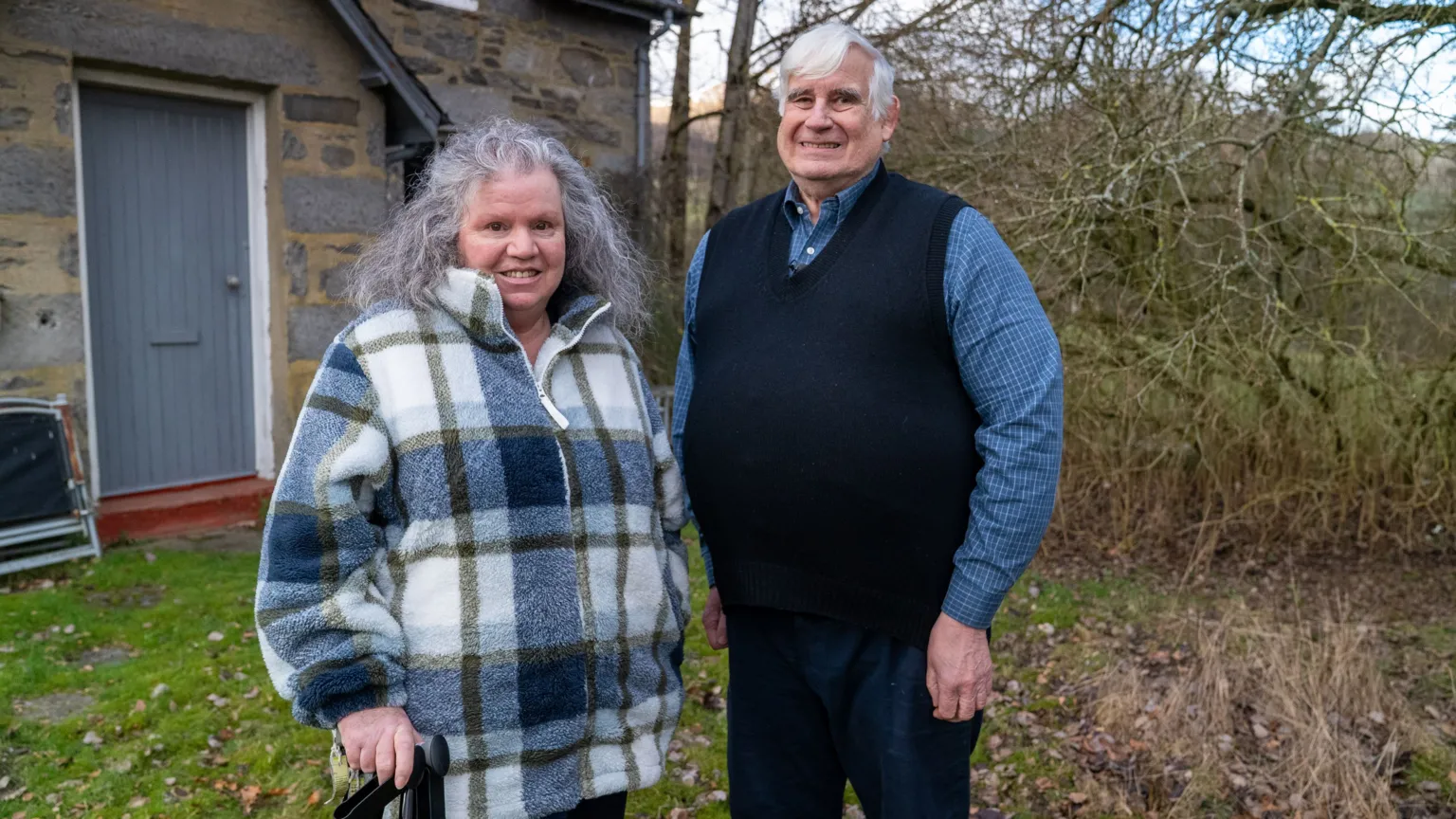 Ann Pearston, a woman with long grey hair wearing a blue, white and gold checked fleece jacket, stands beside Mick North, a man with grey hair in a blue shirt, blue tank top and blue trousers, in a garden. A stone house is behind them and the trees are bare.