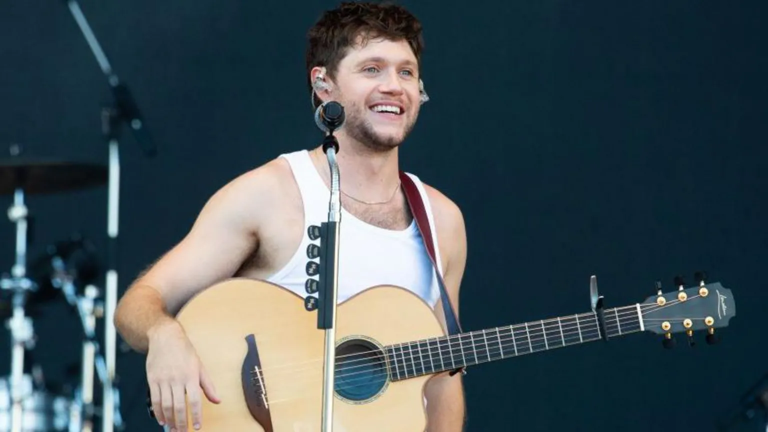 Niall Horan performs on stage at a festival. He's a white man and is standing with a guitar on a strap around his shoulders with his right arm resting on the end of the instrument. He's wearing a white vest and is smiling looking into the distance.