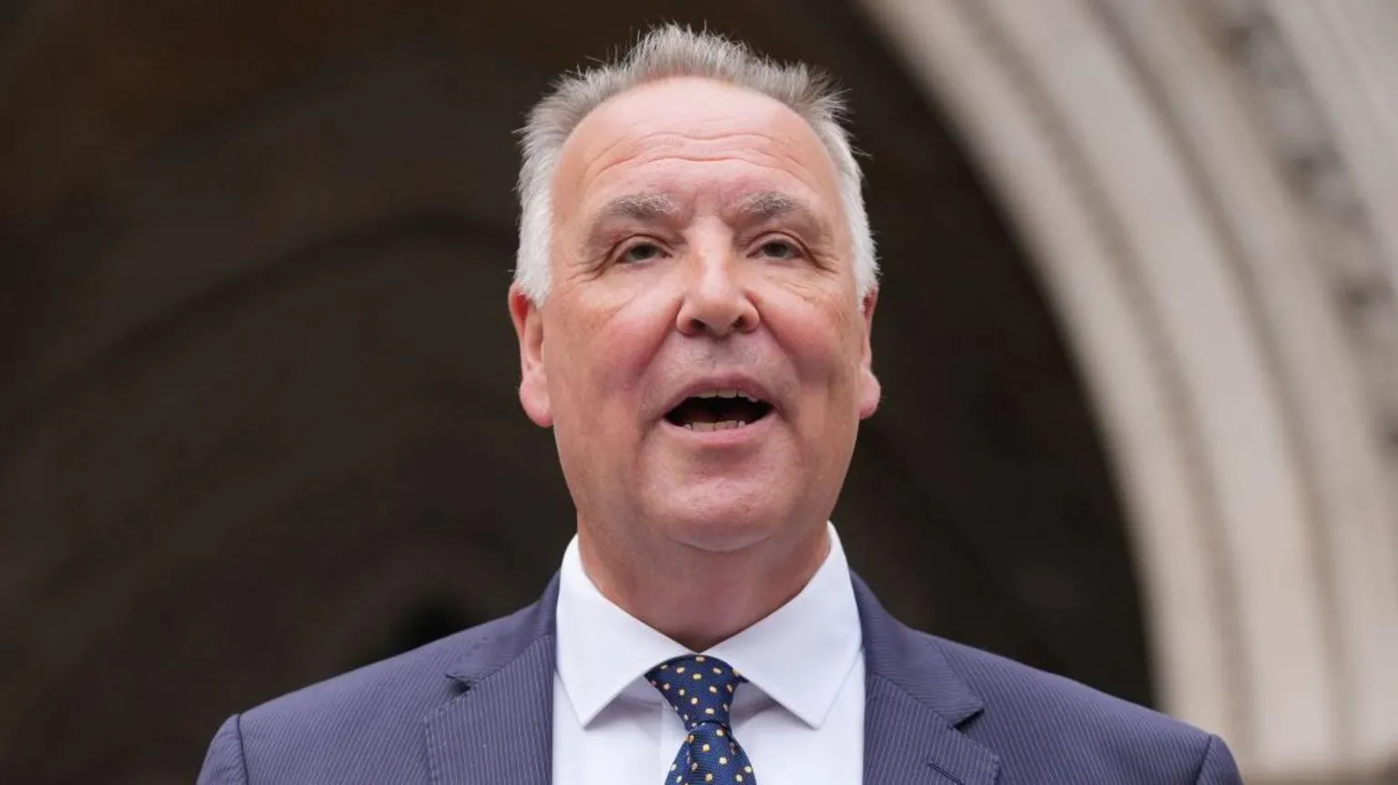  Chris Whitbread has short grey hair and is speaking in front of a cream archway at the High Court. He is wearing a navy suit jacket, white shirt and navy and yellow tie.