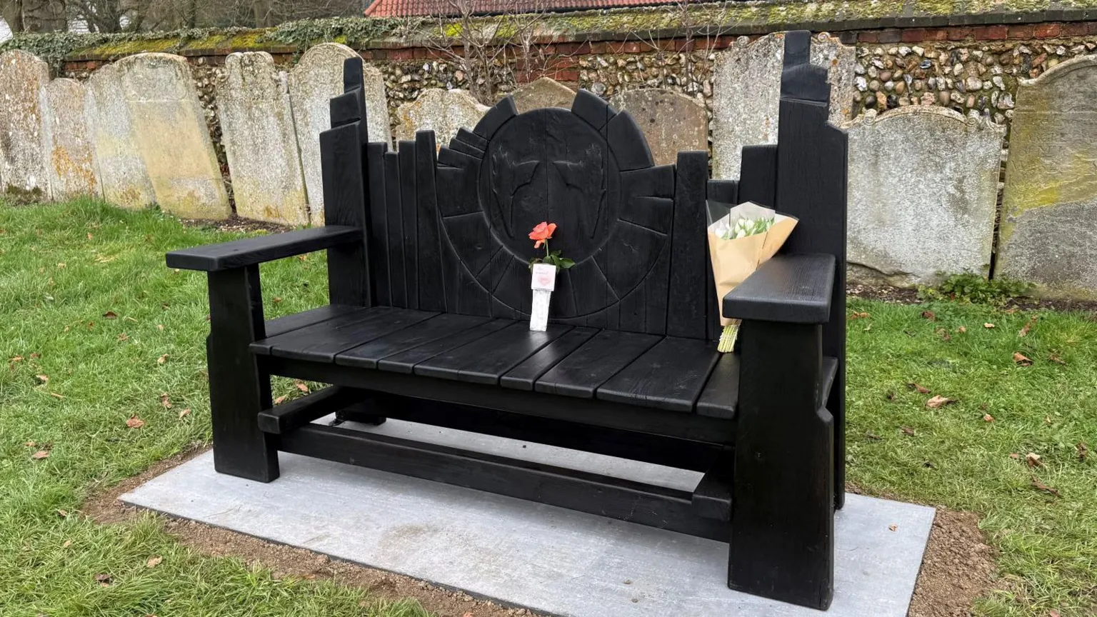 Elliot Deady/BBC Black painted bench with flame carvings and flowers placed on it, on grass with gravestones in the background. 