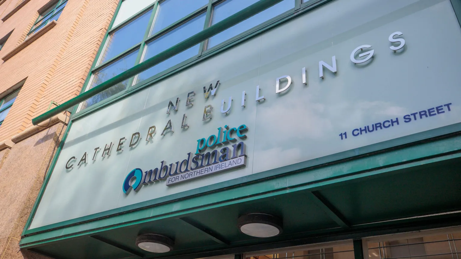 Above the entrance to the police ombudsman's office a sign reads 'NEW CATHEDRAL BUILDINGS: police Ombudsman FOR NORTHERN IRELAND'. The lettering is silver, navy and green, and is set against a background of lighter, pastel green. 