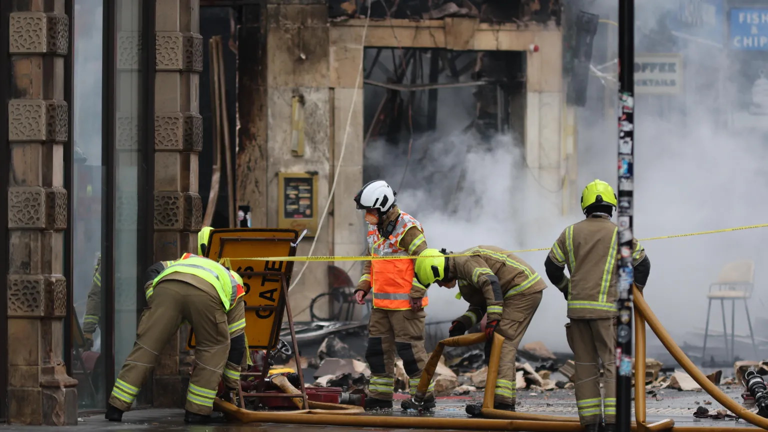  A team of firefighters on the pavement in front of a collapsed building. Rubble lies on the ground and there is smoke in the background.