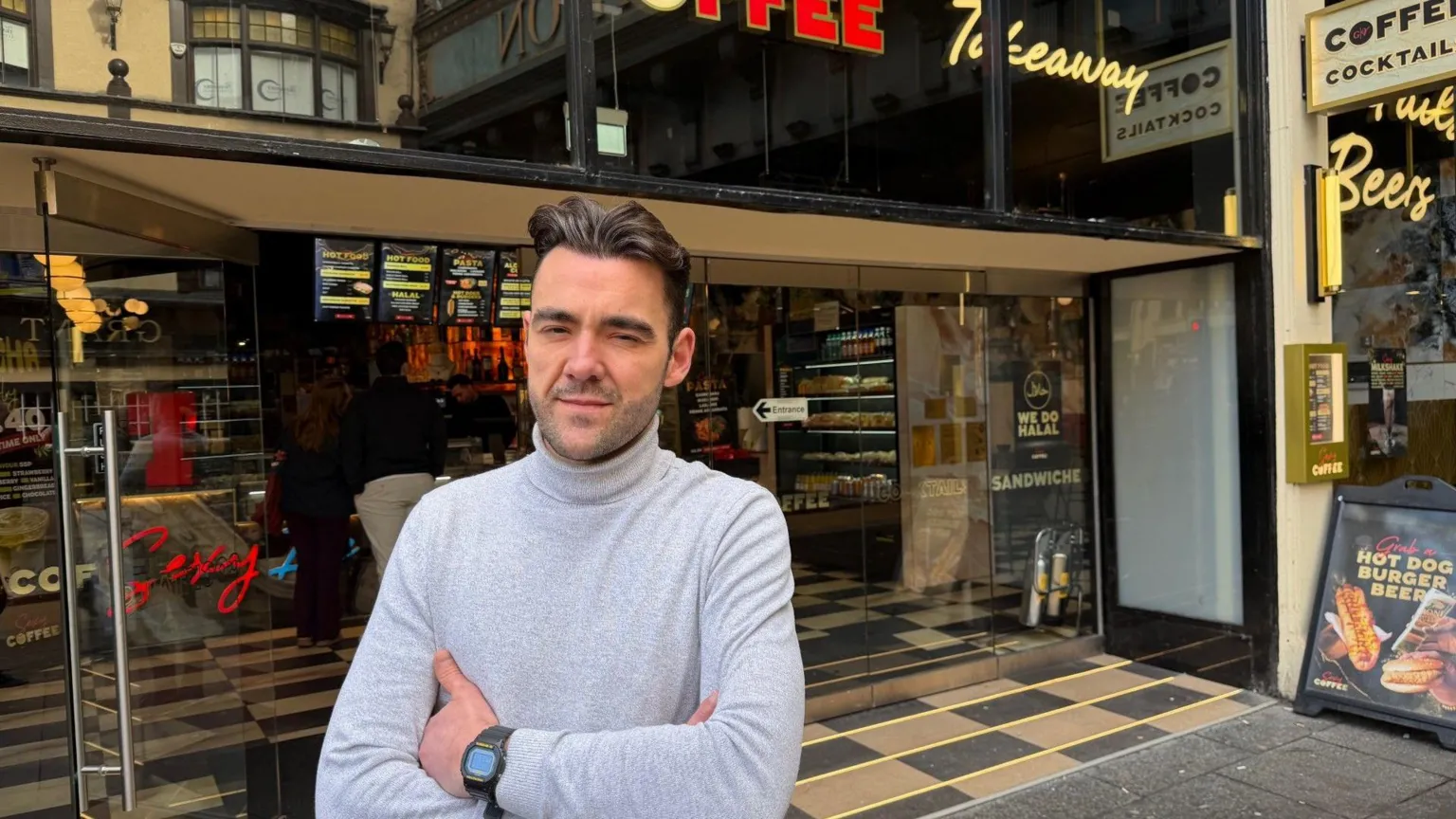 Alessandro Varese A man wearing a grey turtle neck stands in the doorway of a coffee shop with his arms folded