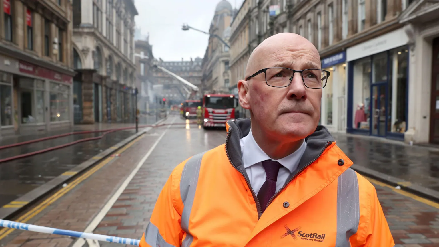  Swinney wearing a bright orange ScotRail high‑visibility jacket stands in front of an emergency cordon on a wet city street, with fire crews and fire engines working in the background amid smoke
