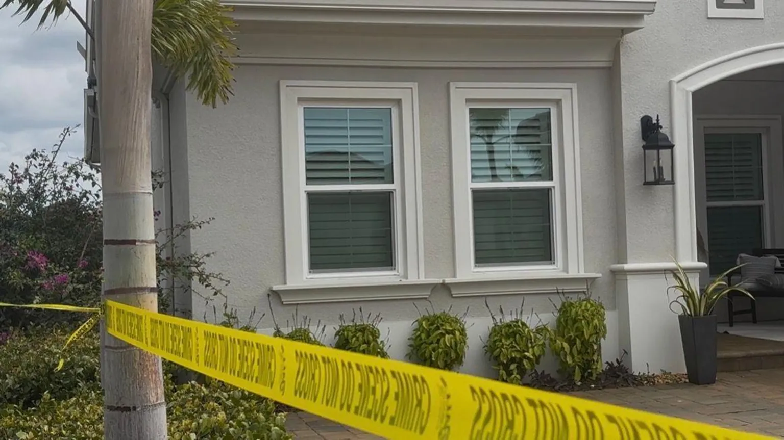 Manatee County Sheriff’s Office A grey/cream Florida home, with a palm tree in the foreground. There is yellow police tape wrapped around the tree. It's a close up picture of the house, showing two front facing windows, with a series of plants running around the front of the house.