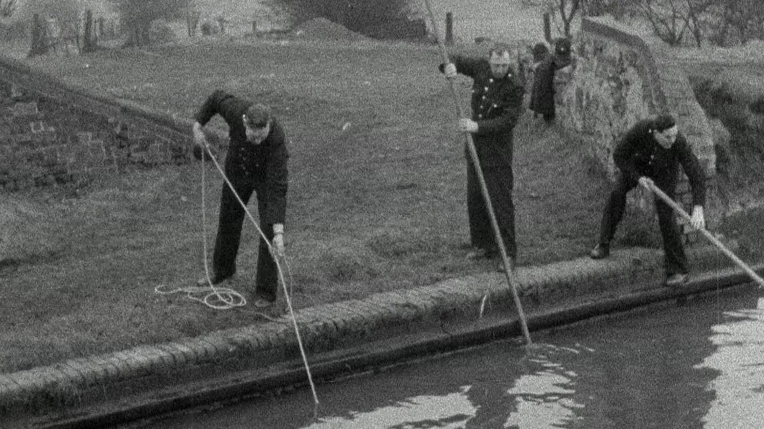A still from black-and-white news footage dating back to the original investigation. It shows three uniformed police officers searching a waterway from the bank with poles and ropes.