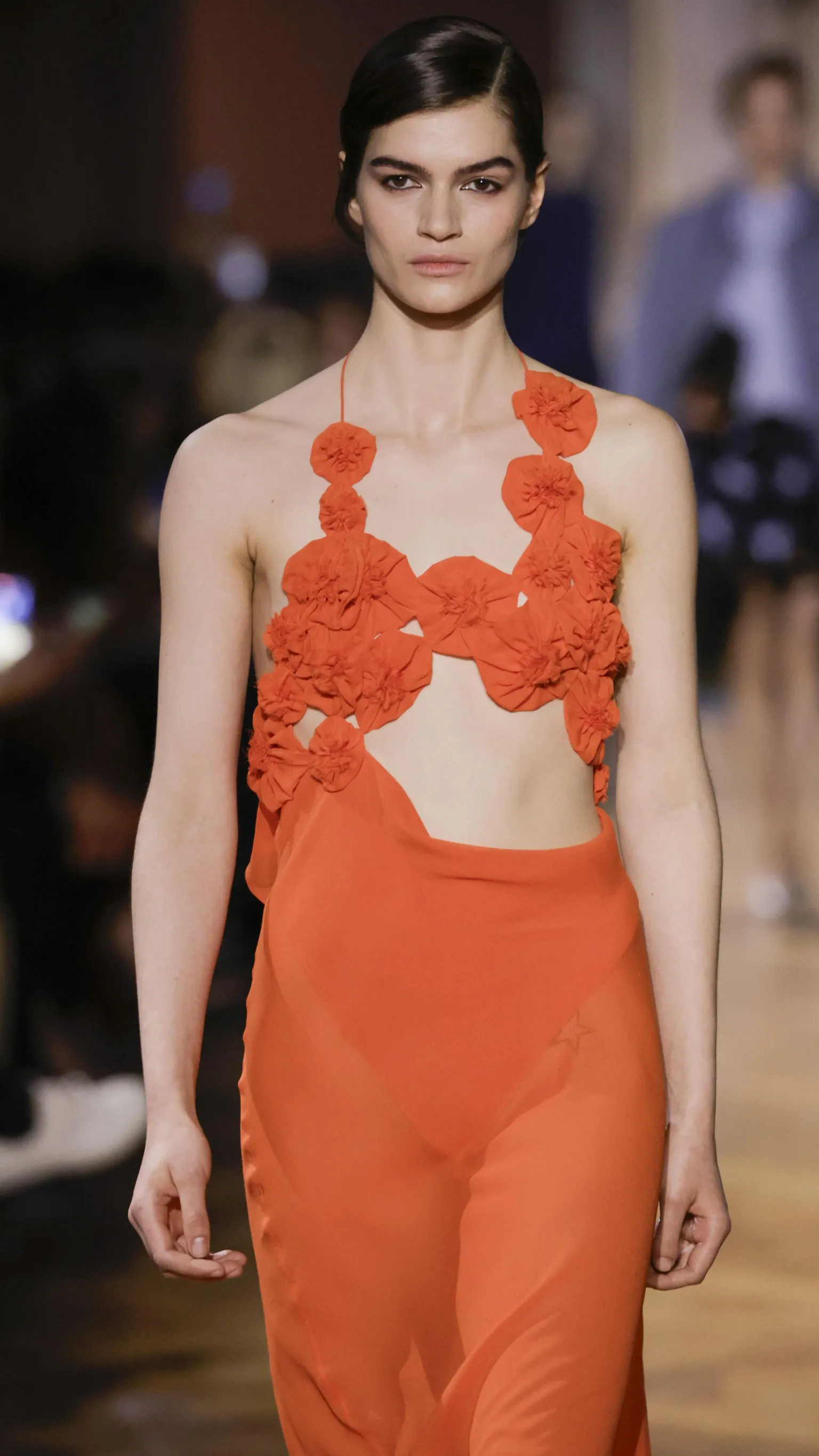  A model wears a coral/red sheer dress with a floral design and a cut-out waist section.