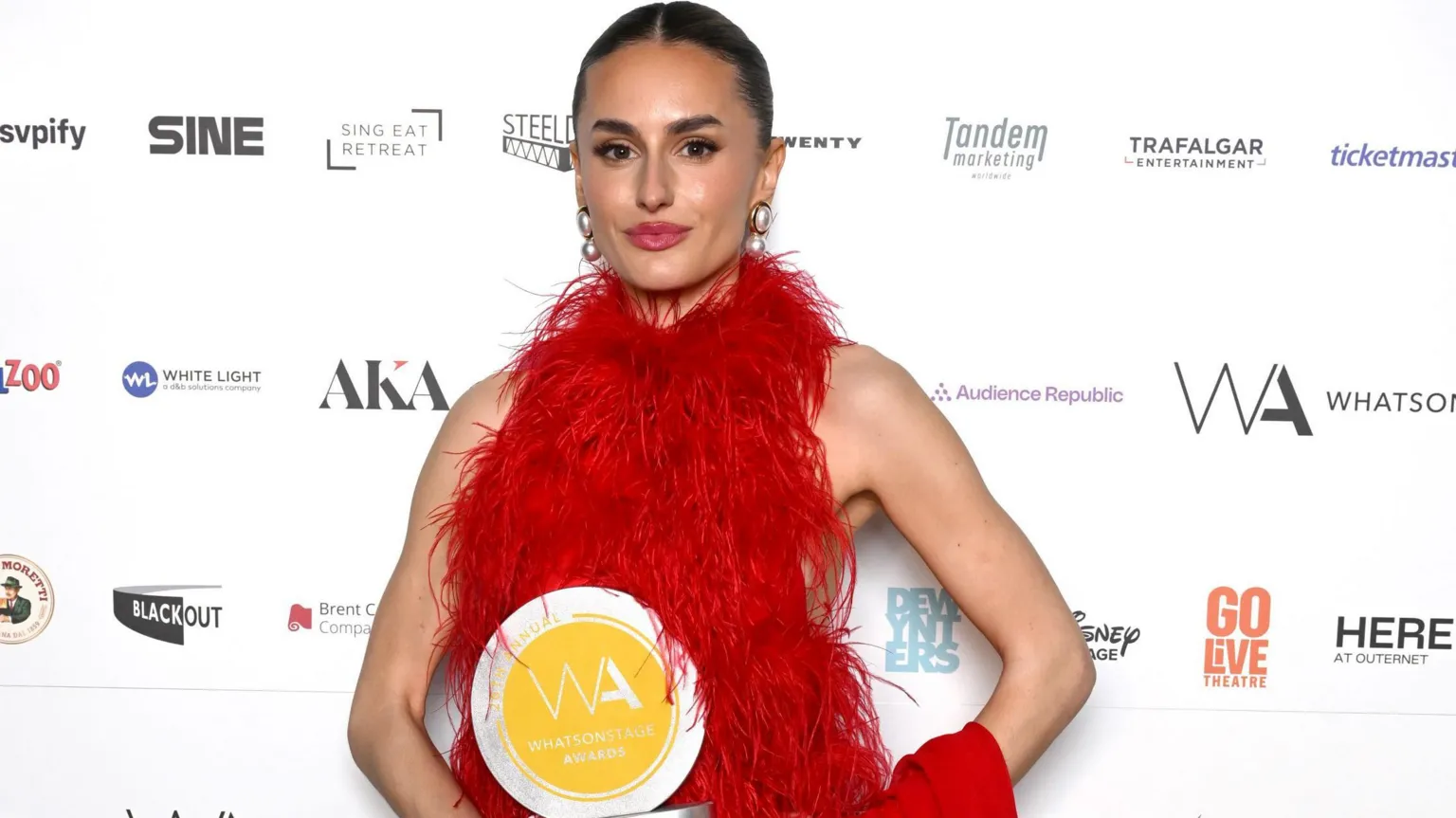  Amber Davies, holding her circular yellow WhatsOnStage award. She has a brunette slick-back bun, wearing a red dress and big pearl earrings. She stands in front of a branded white wall with sponsor logos.