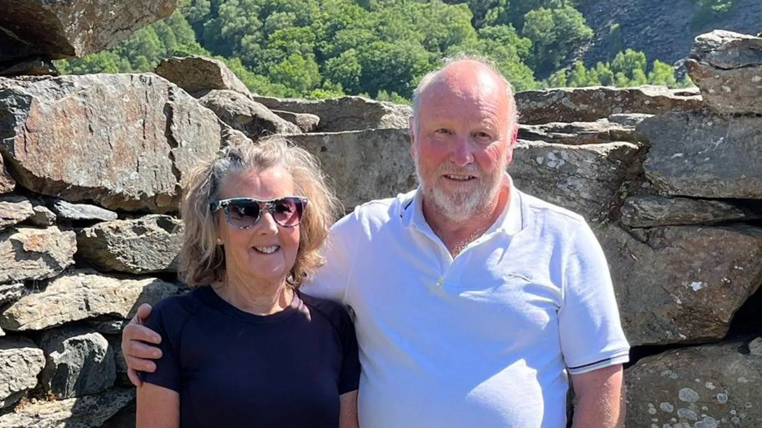 Christopher Lavis Elizabeth and Christopher Lavis in short sleeve shirts and shorts and next to an old stone wall and stone sculpture of a large dog.