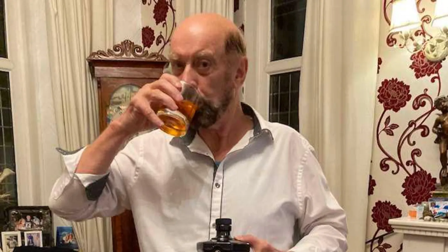 Facebook John Sexton Sr is a bald man with a beard. He is in an open necked white shirt, presenting and drinking from a bottle of whisky. It is a large glass of whisky. The room behind him has thick cream curtains, burgundy and cream wallpaper and an antique clock.
