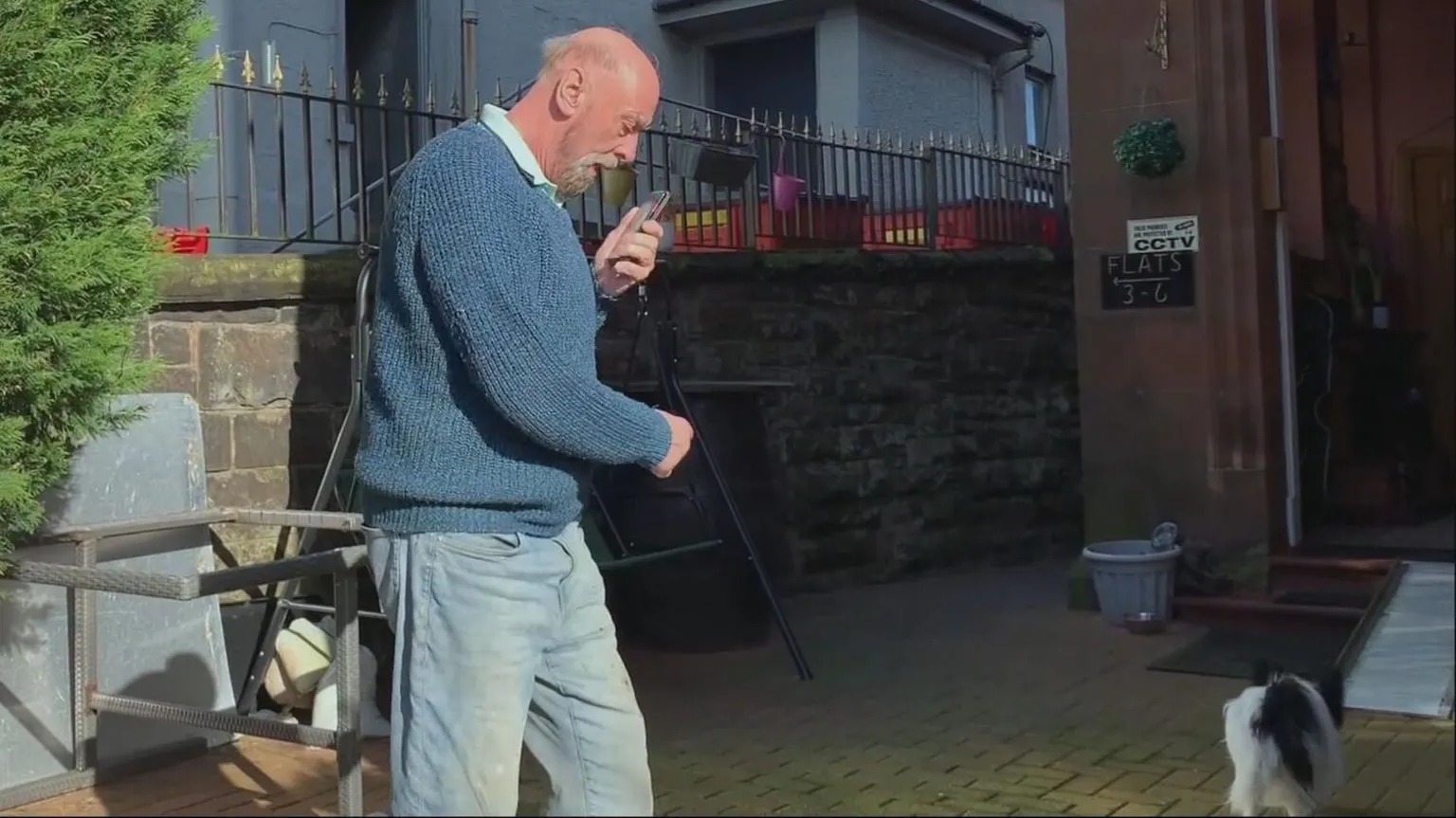 John Sexton Sr in a blue jumper and jeans is speaking into a phone and walking away from the photographer in the garden of a large red sandstone house. Garden furniture can be seen stacked and a small black and white dog is walking away too.
