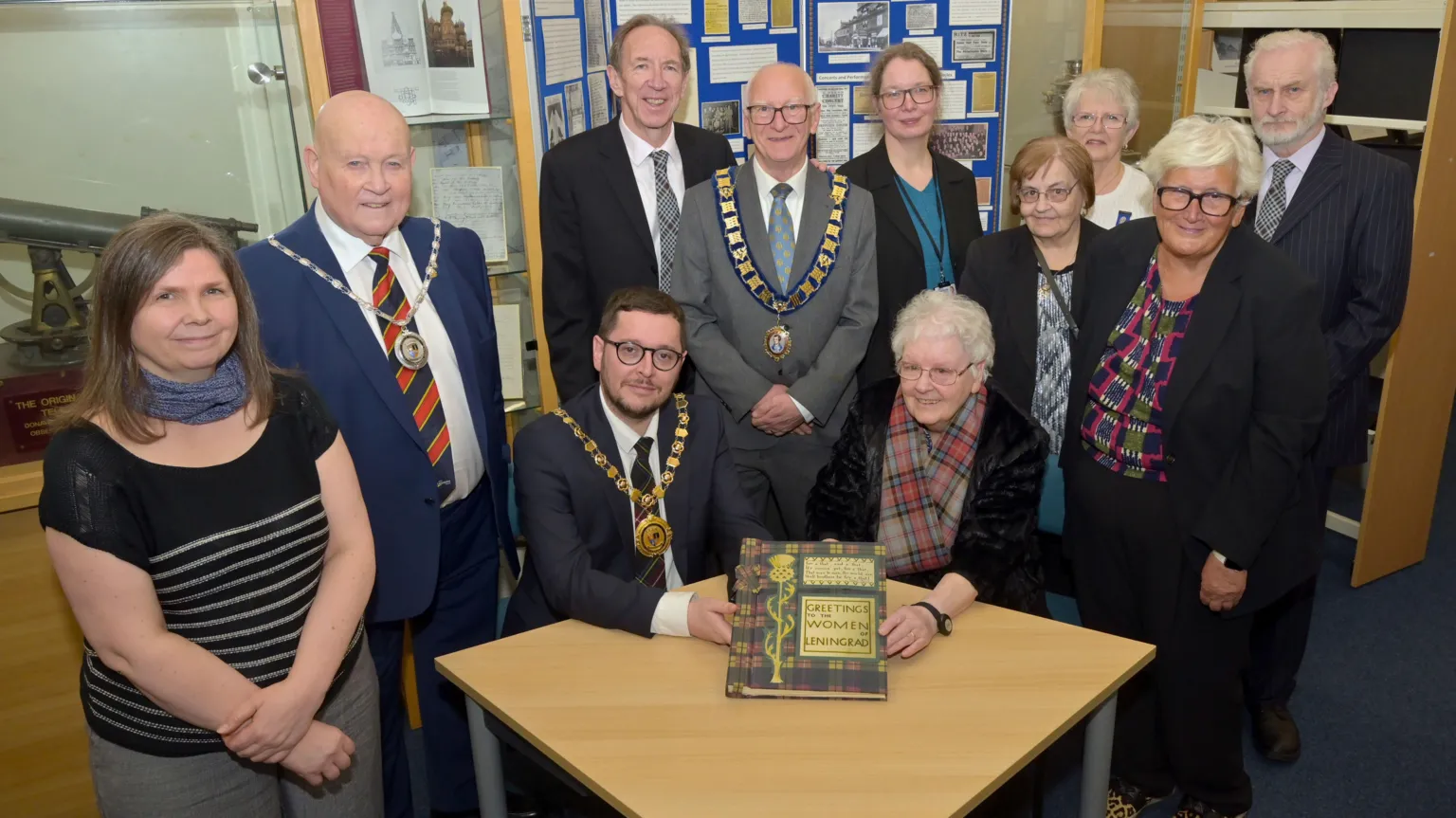 North Lanarkshire Council Several people surrounding a table, with two people sitting at the table. A woman - Helen Morrison - is sitting at the table with a tartan scarf and white hair, holding a a book with a tartan cover and titled Greetings to the women of Leningrad on the front. Sitting alongside her is a man in a suit with short dark hair, glasses and a lord provost's chain.