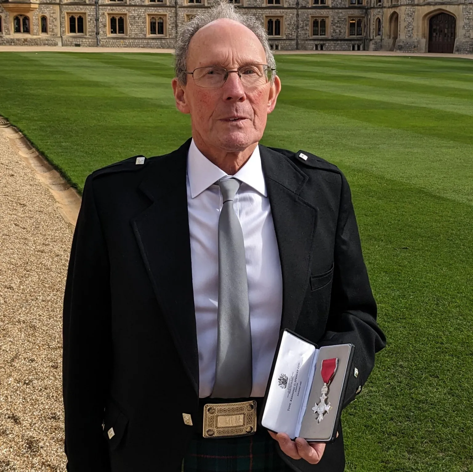 Malcolm Kirby A man wearing a kilt and holding a medal in front of a mowed lawn and grand castle.