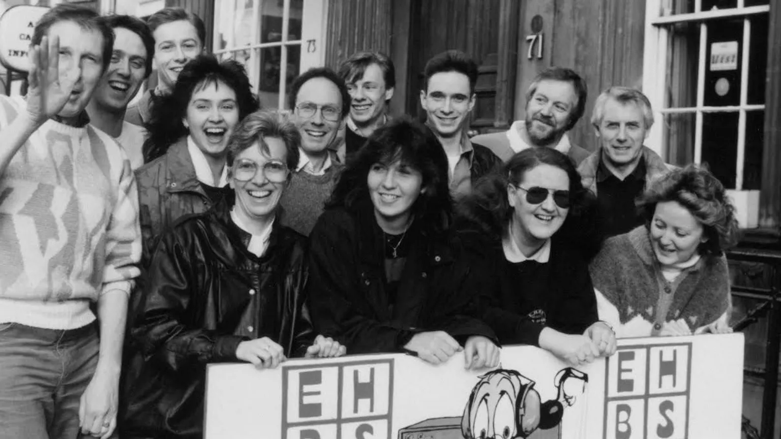 Malcolm Kirby A black and white image of the hospital radio volunteers smiling and holding a sign