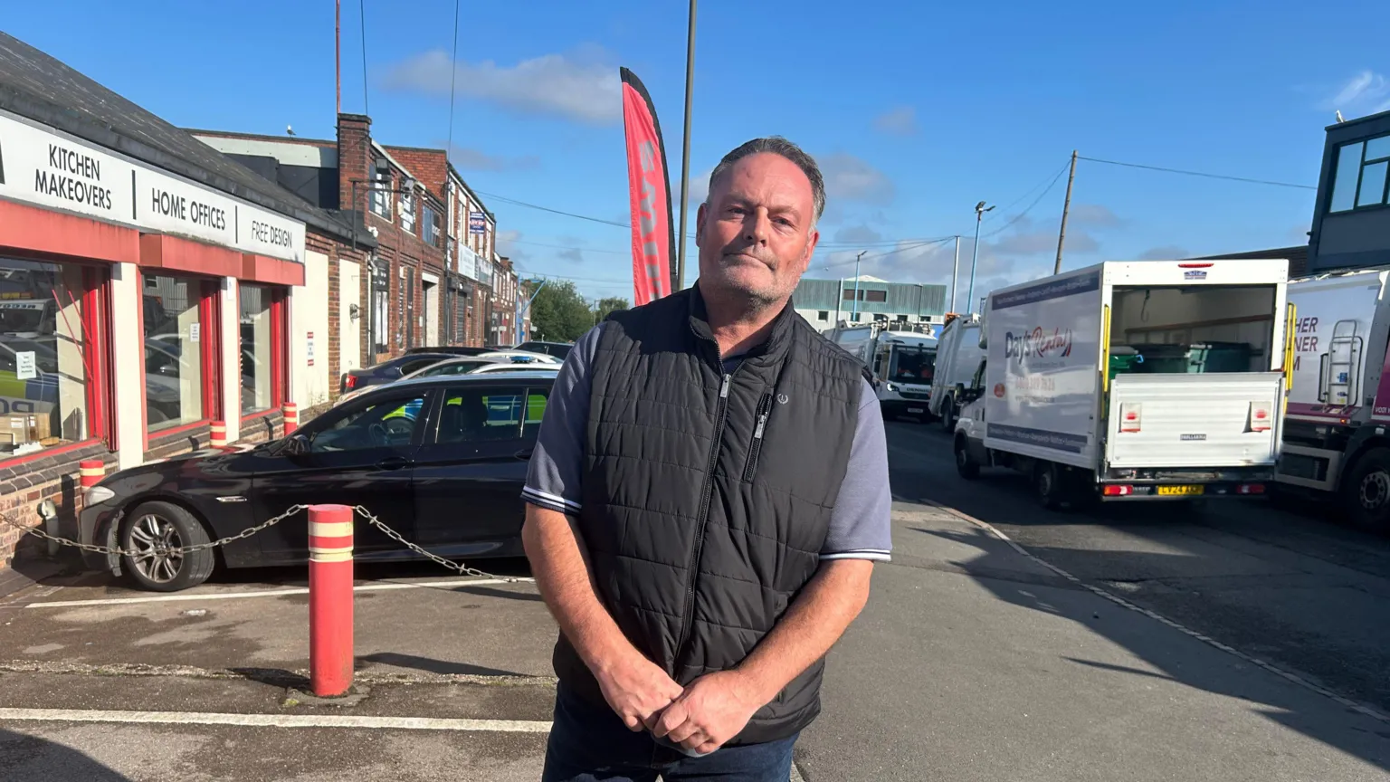 Derek Roberts in a blue polo shirt and black gilet is looking at the camera. In the background are shops and industrial buildings, and several cars are parked on the left, including a police car. Lorries are on the adjacent road.