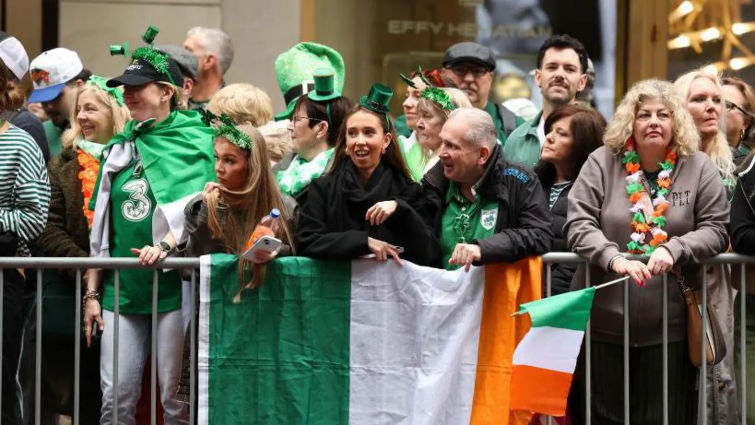  A number of people with green hats and head pieces on, and holding Irish flags as they stand behind a barrier watching the St Patrick's Day parade in New York.