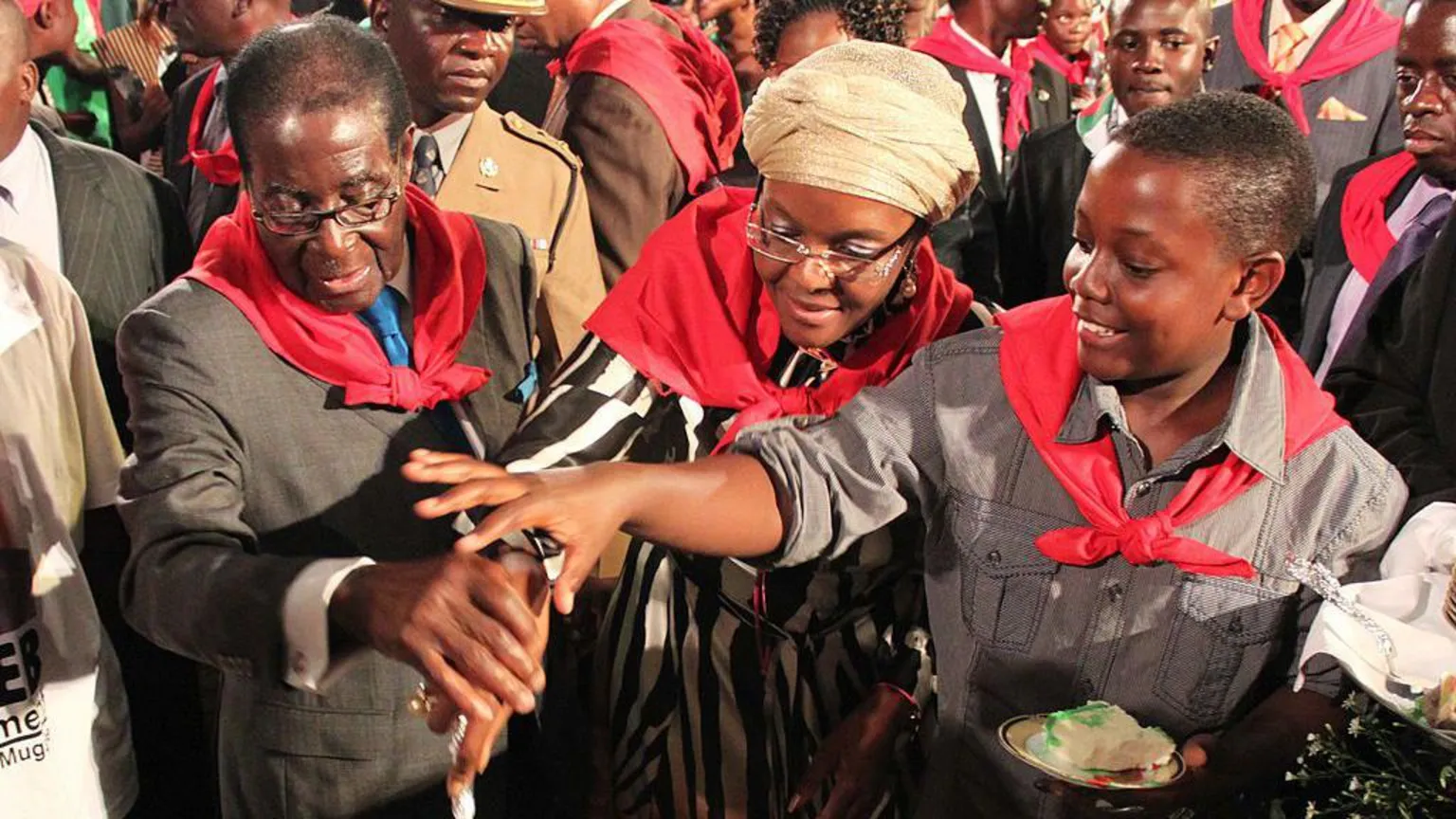Gallo Images/ Robert, Grace and Bellarmine Mugabe are standing together, smiling and wearing red neck scarves as they jointly cut a cake.