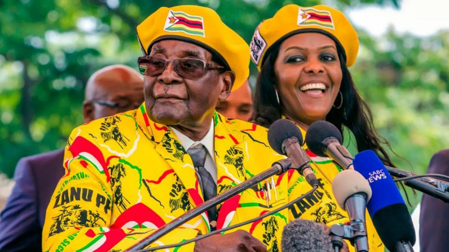 AFP via Grace and Robert Mugabe, dressed in bright yellow Zanu-PF clothes and berets stand next to each other at some microphones.
