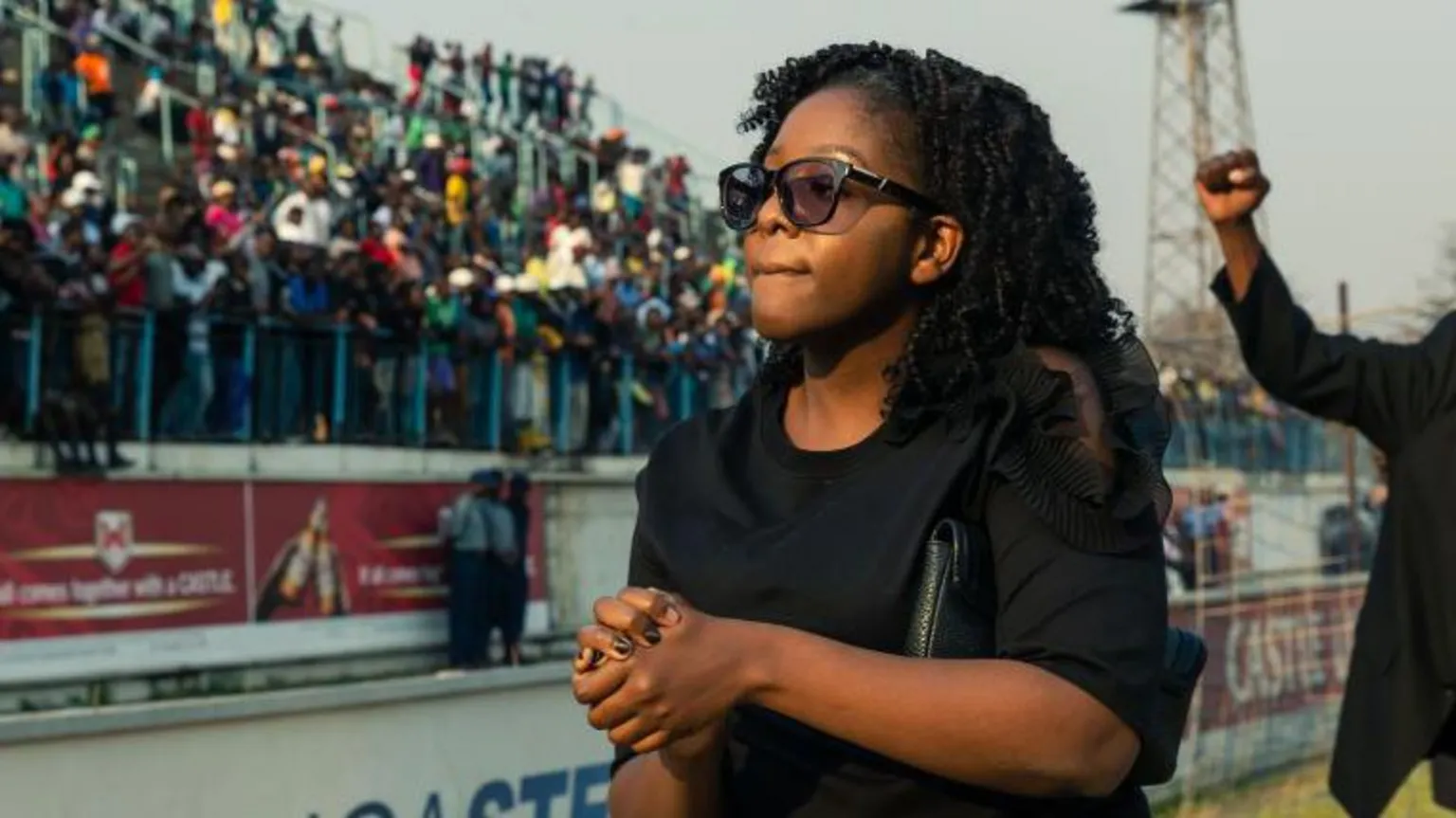 AFP via Bona Mugabe is wearing sunglasses and dressed in black has her hands clasped in front of her. A crowd of people in a stadium stand can be seen out of focus behind her.