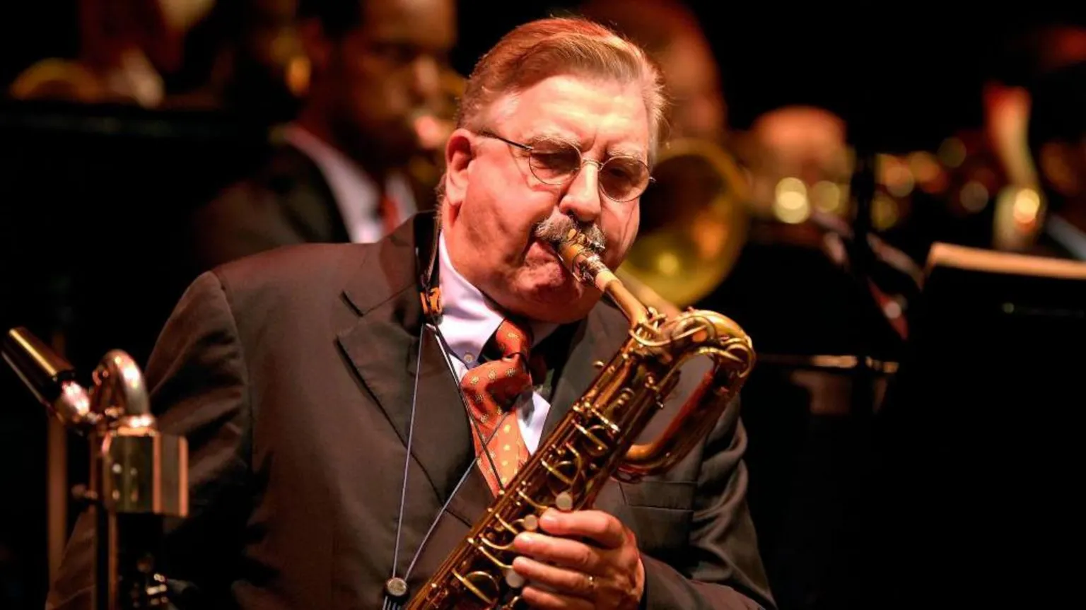 Joe Temperley, an elderly gentleman in brown suit, blue shirt and red tie, plays the saxophone. Other band members can be seen behind him.