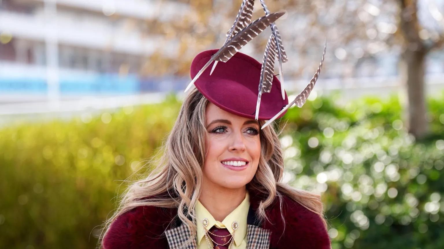 A young woman with wavy blonde hair smiles as she poses for the camera at Ladies Day at the Cheltenham Festival 2026. She is wearing a plum red berry-style beret with feathers attached to it, and a jacket that matches her hat