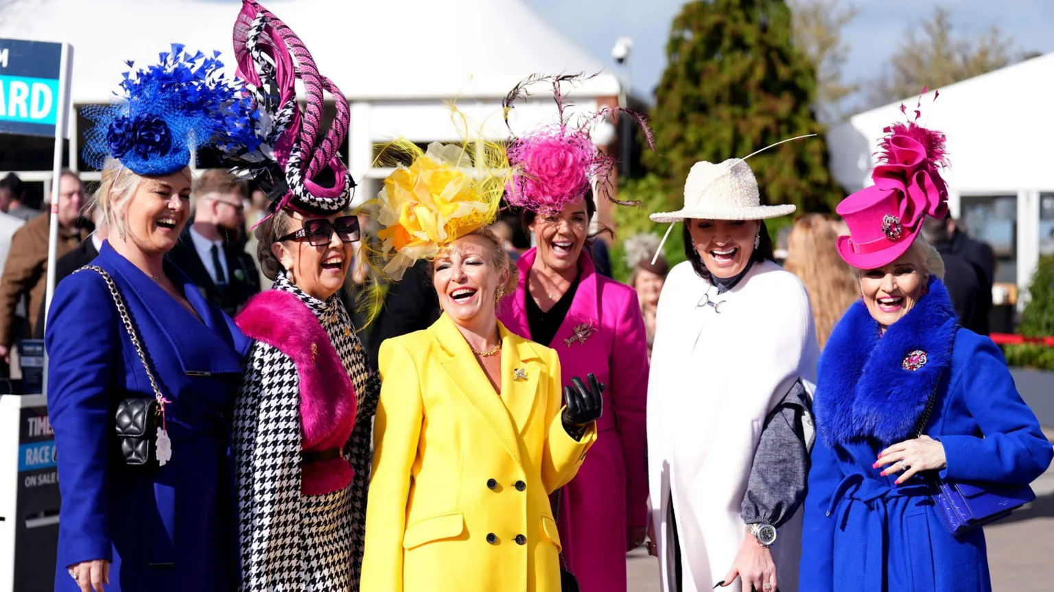 A line of women laugh as they pose for the camera at the start of Ladies' Day at the 2026 Cheltenham Festival. Some of them have bright yellow or blue outfits on, and all of them are wearing bright fascinators on their heads