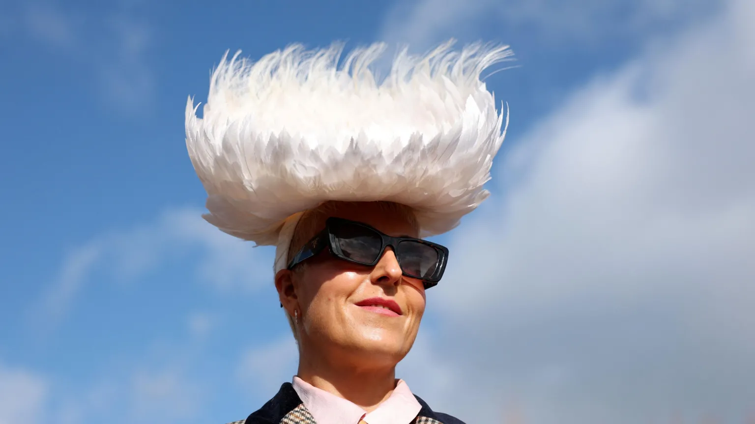 A woman with large sunglasses and a white, fluffy hat poses for the camera as Ladies' Day gets under way at the Cheltenham Festival. Behind her is a mixture of blue sky and cloud