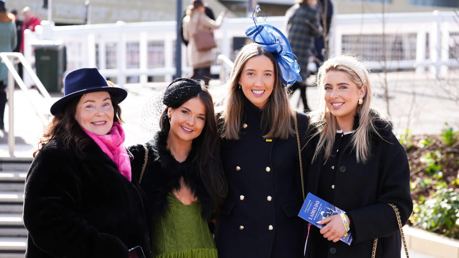 Four women, three of them young and one slightly older, smile as they stand arm in arm at the Cheltenham Festival. They are all wearing smart dark jackets and hats or fascinators