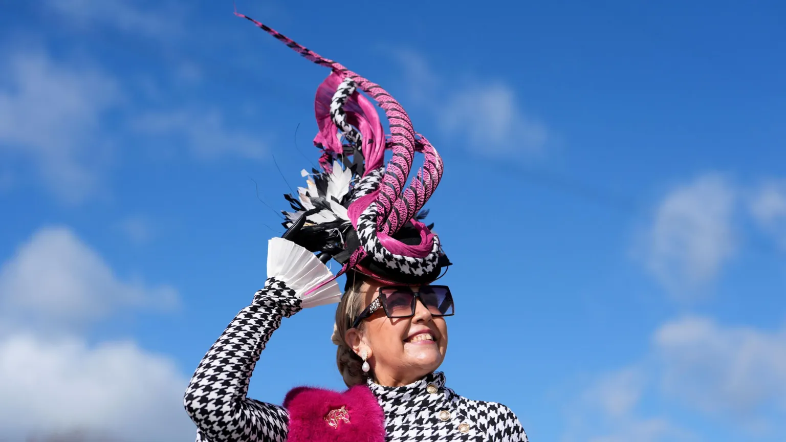 A woman with a black and white checked jacket holds onto a dramatic piece of headgear at the second day of the 2026 Cheltenham Festival. The hat is made of long, curling pieces of black and purple material