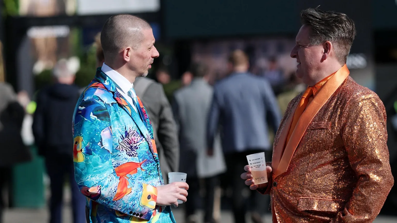 Two men face each other as they chat while holding drinks. One has a shiny orange formal suit on, while the other is wearing a light blue suit with colourful emblems on it