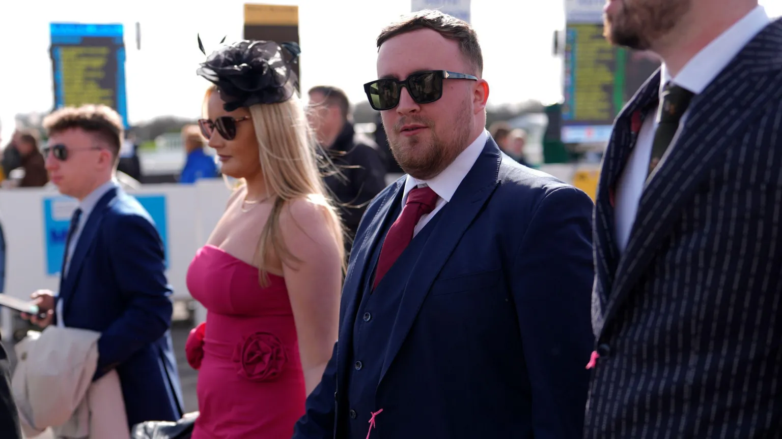 Darts star Luke Littler walks into the Cheltenham Festival, with his other guests either side. He is wearing a dark blue suit and waistcoat, a white shirt and dark pink tie and sunglasses