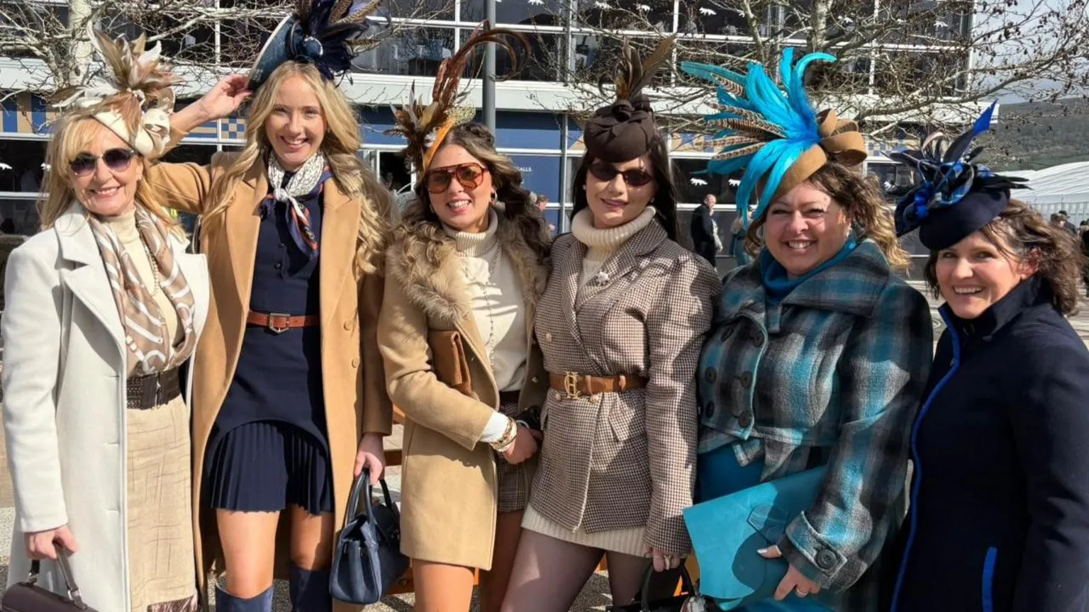 Six women of various ages all wearing fascinators with feather details smile at the camera. They are wearing smart coats and jumpers and skirts or dresses.
