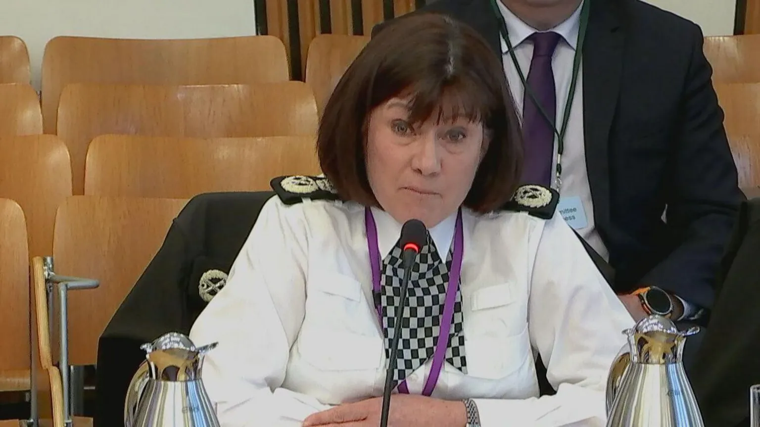 Scottish Parliament TV Chief constable Jo Farrell wearing a white shirt with a black and white checked necktie and black patches on the shoulders sitting behind a microphone at the Scottish Parliament.