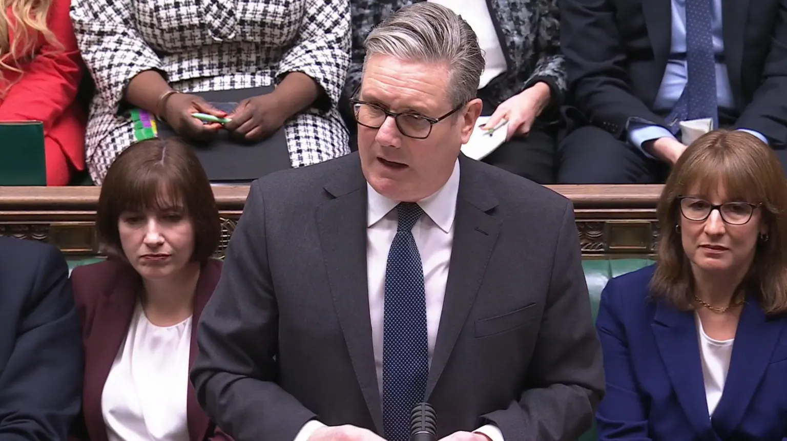  Sir Keir Starmer, a man with grey hair and glasses wearing a dark grey suit and blue tie, speaks while standing in front of green leather-seated benches occupied by members of Parliament in the House of Commons.