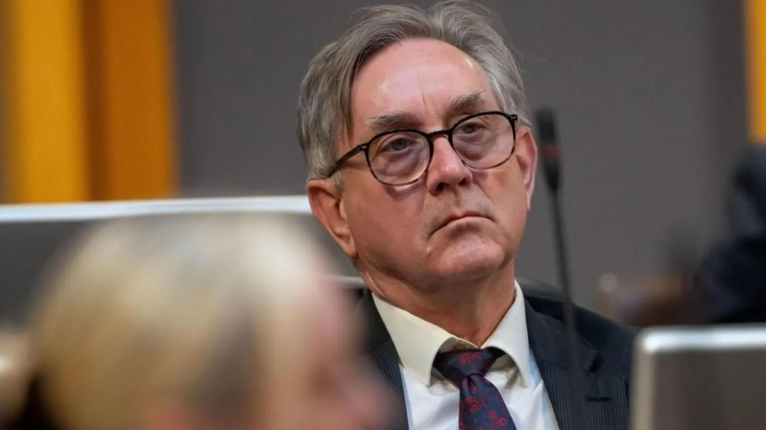 Senedd Cymru Mick Antoniw, a man with grey hair and glasses wearing a navy suit and tie, is sat down in a large room.