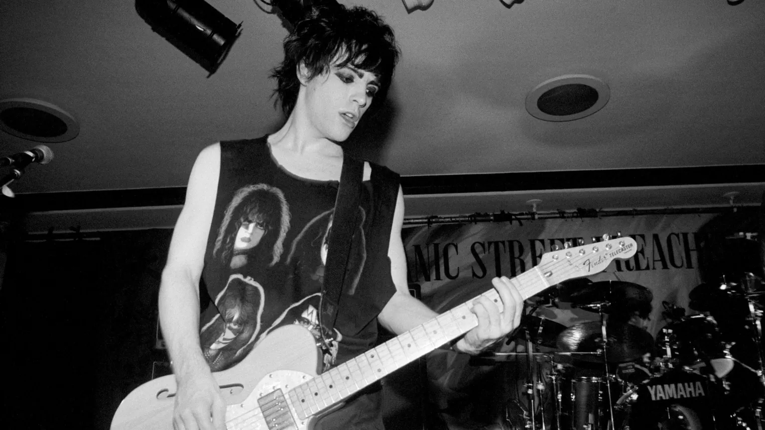 Black and white picture of Edwards. He plays an electric guitar. He stands in front of a drum kit. Wears a vest top with the pictures of glam rockers featured